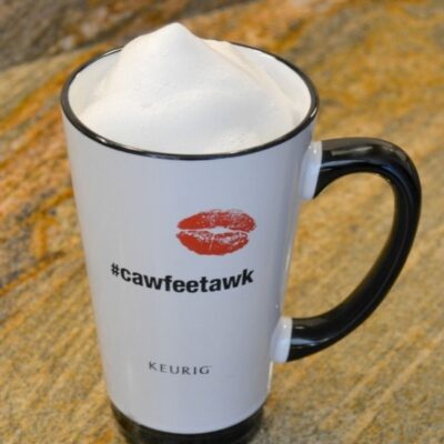 White mug with a black handle, filled with frothy foam, sits on a brown countertop. Perfect for easy homemade skinny lattes, the mug reads "#cawfeetawk" with a red lipstick print above the text.