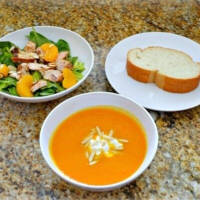 A bowl of Ginger Carrot Soup with a dollop of cream, paired with an Asian Chicken Salad featuring greens and orange slices, plus two slices of bread on separate plates.