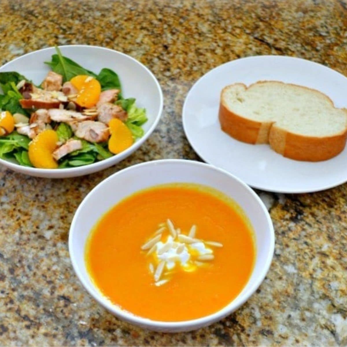 A bowl of Ginger Carrot Soup with a dollop of cream, paired with an Asian Chicken Salad featuring greens and orange slices, plus two slices of bread on separate plates.