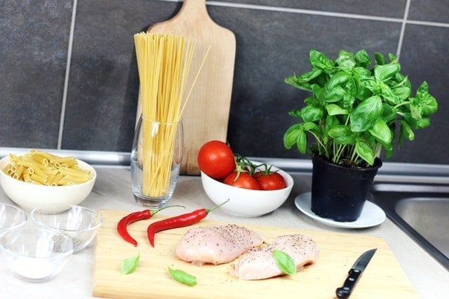 pasta, vegetables, raw chicken on a cutting board next to a knife and a plant