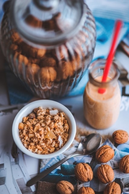 granola in a white bowl next to a jar of walnuts, walnuts on a table next to spoon, and a jar of a drink with a straw in it