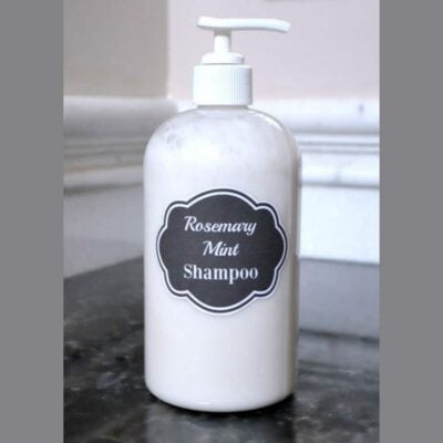 A clear pump bottle labeled "Rosemary Mint Shampoo" sits on a dark countertop against a light wall, showcasing fresh homemade rosemary mint shampoo.