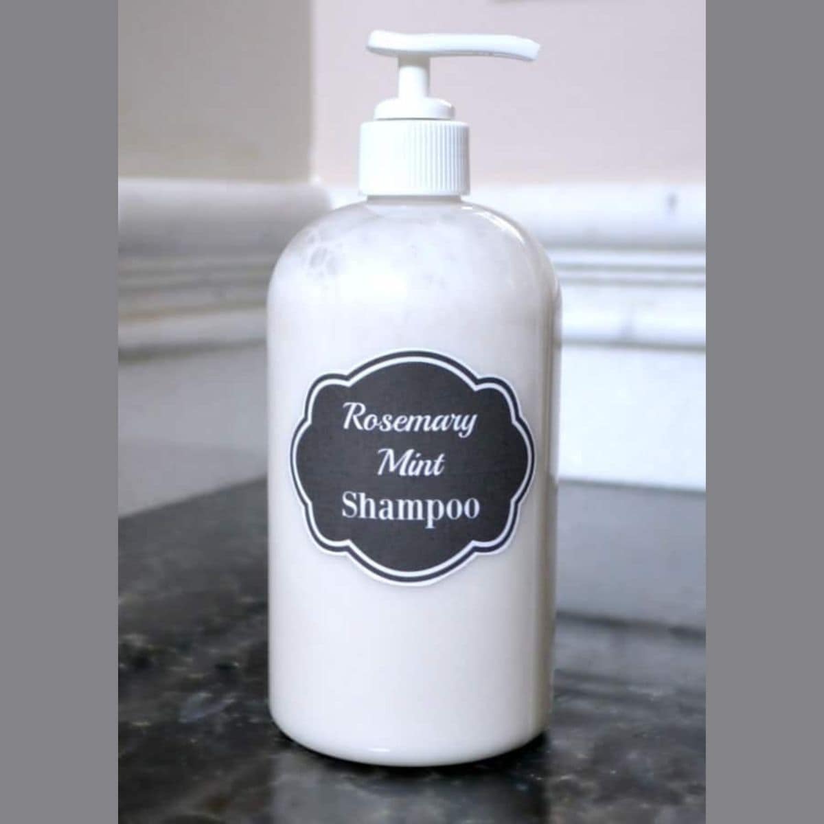 A clear pump bottle labeled "Rosemary Mint Shampoo" sits on a dark countertop against a light wall, showcasing fresh homemade rosemary mint shampoo.