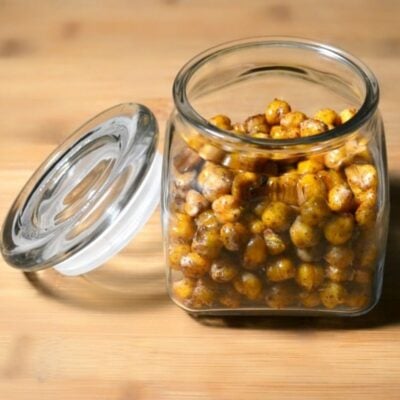 A glass jar filled with seasoned roasted chickpeas sits on a wooden surface with its lid resting nearby, perfect for trying out your favorite honey roasted chickpea recipe.