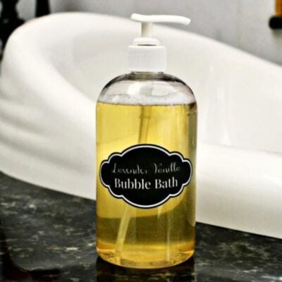 Clear bottle with pump dispenser labeled "Lavender Vanilla Bubble Bath," a natural homemade bubble bath, sits on a dark countertop in front of a white bathtub.