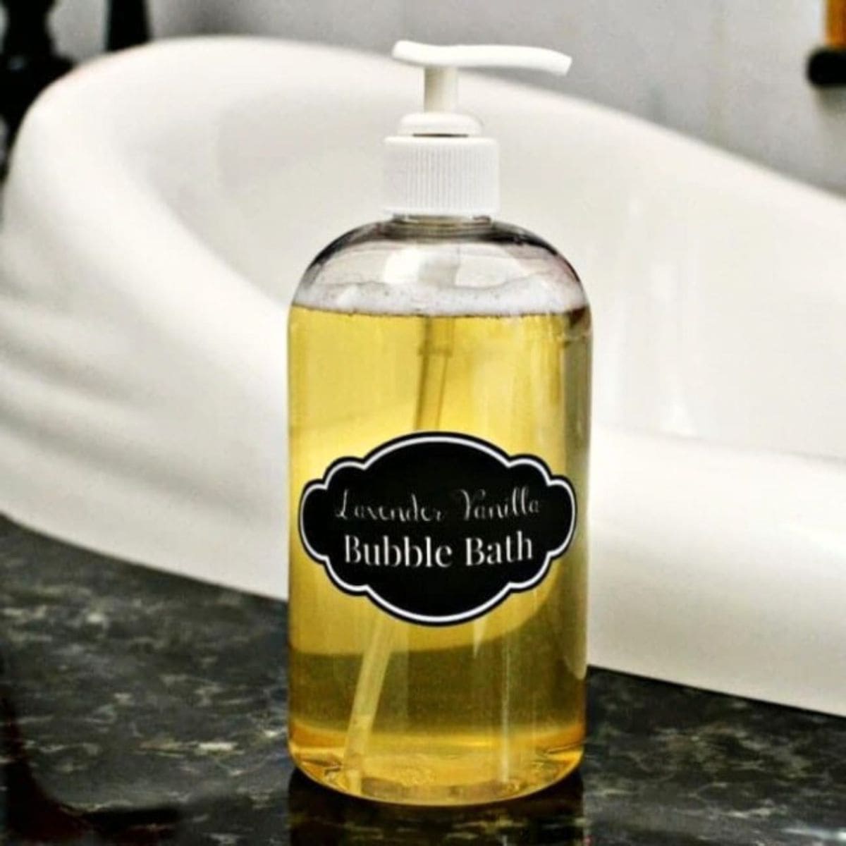 Clear bottle with pump dispenser labeled "Lavender Vanilla Bubble Bath," a natural homemade bubble bath, sits on a dark countertop in front of a white bathtub.