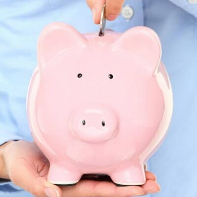 A person in a blue shirt holds a pink ceramic piggy bank with one hand and inserts a coin with the other, symbolizing commitment to a family savings plan.