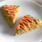 Two triangular carrot cake scones rest on a white plate, each adorned with orange and green icing that charmingly resembles carrots.