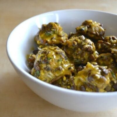 A white bowl filled with several tangy vegan meatballs coated in a yellow sauce, placed on a light wooden surface.