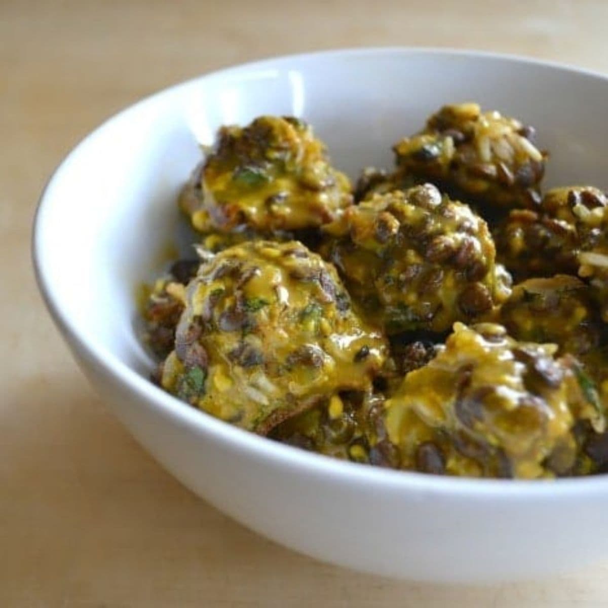 A white bowl filled with several tangy vegan meatballs coated in a yellow sauce, placed on a light wooden surface.