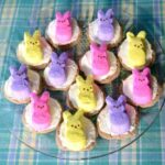 A glass plate brimming with Easter cake mix cookies topped with white frosting and marshmallow bunnies in pink, purple, and yellow sits on a blue plaid tablecloth.