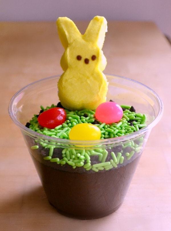 chocolate pudding in a cup with sprinkles, round candy, and a yellow peep on top, on a brown table