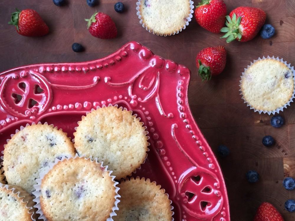 Strawberry Blueberry Muffins in a red dish next to more muffins, strawberries, and blueberries on a brown table