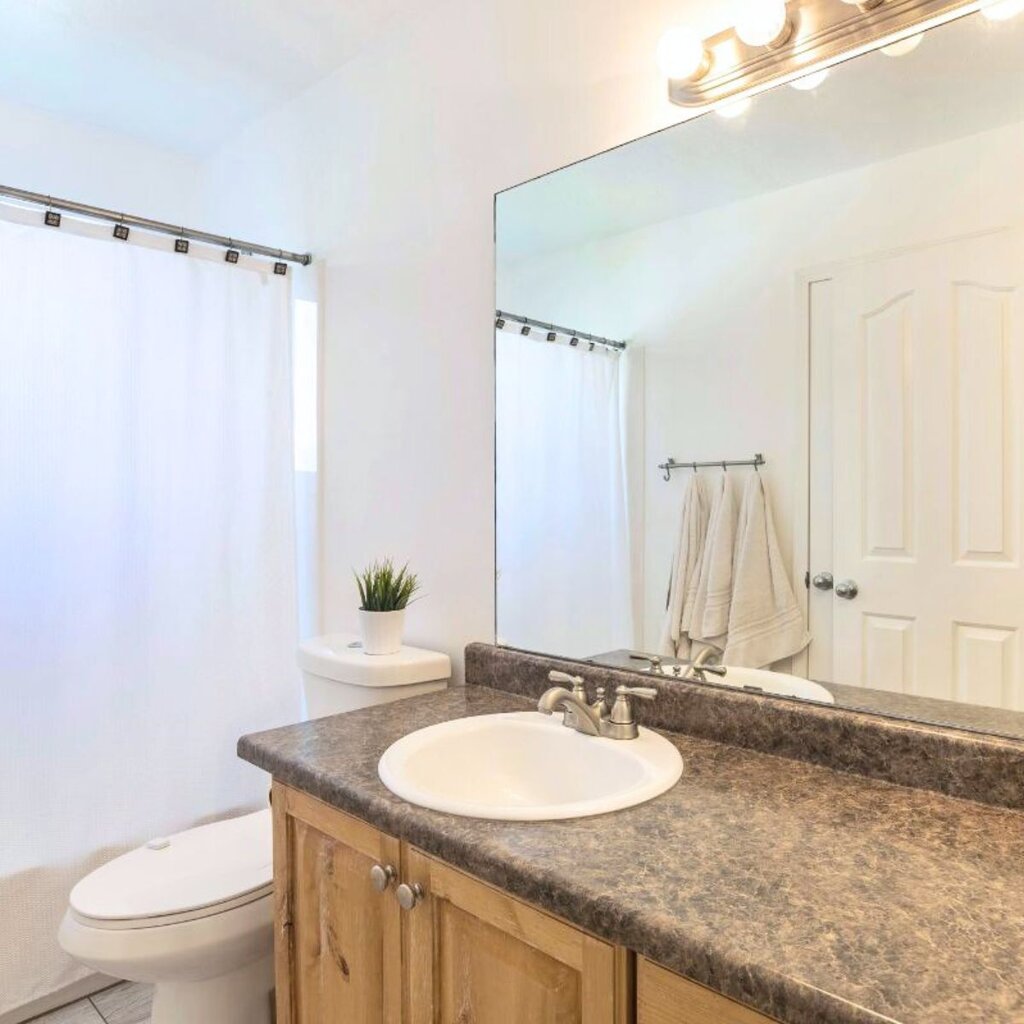 A clean bathroom with a brown countertop, white sink, wooden cabinet, toilet, potted plant, towels on a rack, large mirror, and shower with a white curtain—perfect for making a small space seem bigger.