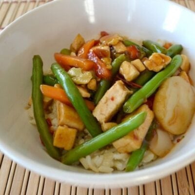 A white bowl filled with rice, tofu cubes, green beans, carrots, water chestnuts, and mixed vegetables in a savory brown sauce—a delicious tofu teriyaki bowl.