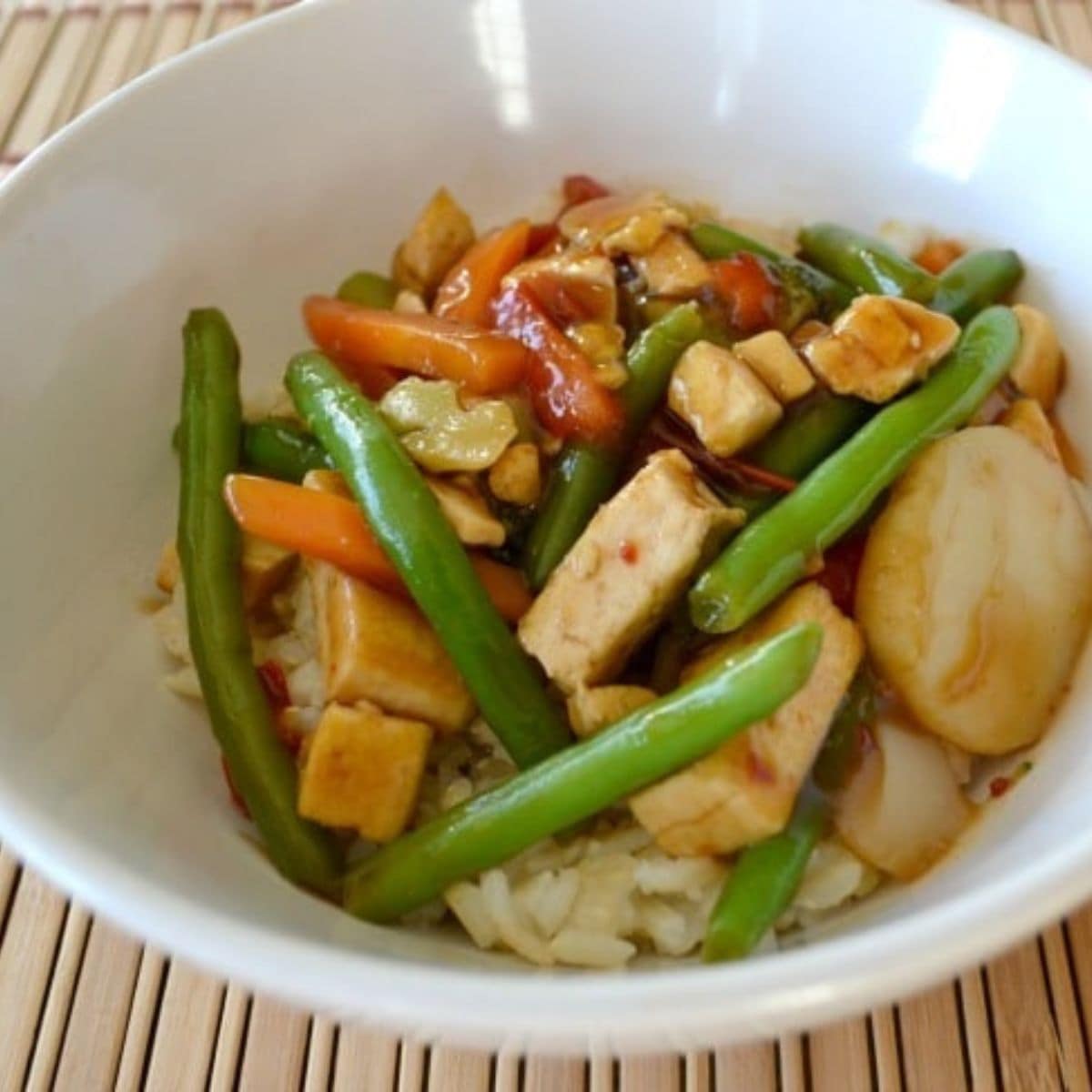 A white bowl filled with rice, tofu cubes, green beans, carrots, water chestnuts, and mixed vegetables in a savory brown sauce—a delicious tofu teriyaki bowl.