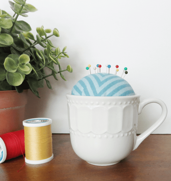 teacup pin cushion next to two spools of thread and a plant on a wood table with a white background