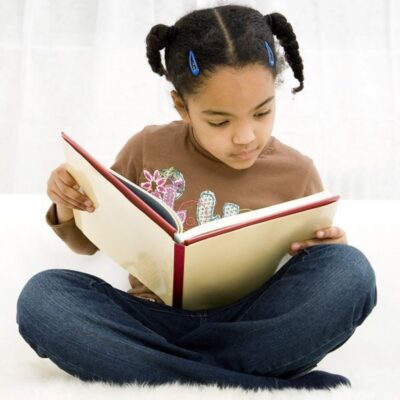 A young girl with braids sits cross-legged and reads a large book, focused on the pages, determined to stop summer learning loss.