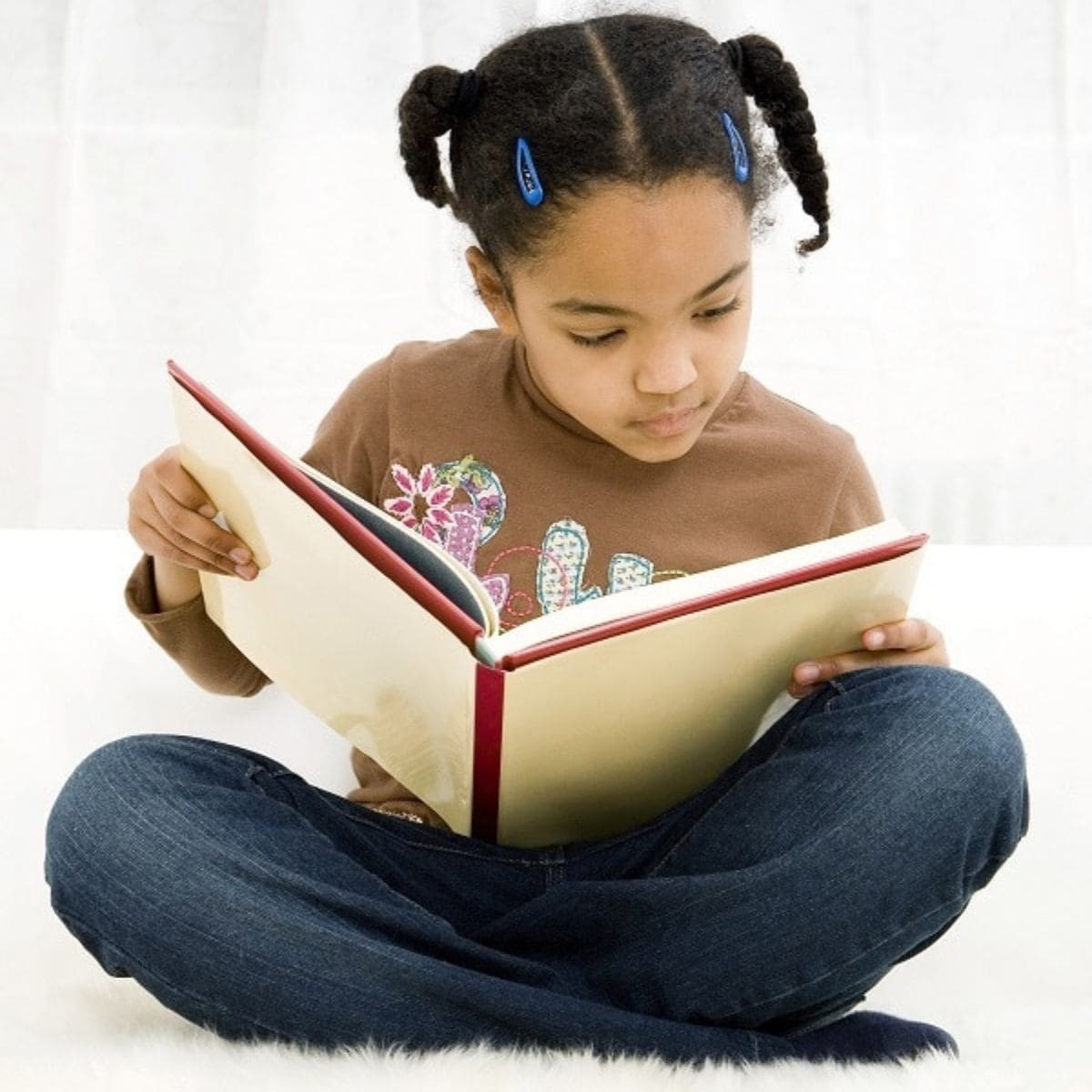 A young girl with braids sits cross-legged and reads a large book, focused on the pages, determined to stop summer learning loss.