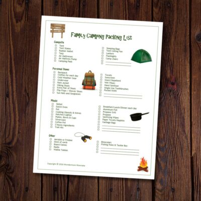 A printed family camping packing list with categorized checkboxes for campfire, personal items, meals, and other essentials is placed on a wooden surface.