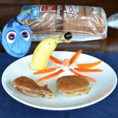 A plate with creative food art: a banana snail with an olive eye, an orange octopus, two fish-shaped sandwiches, and a loaf of bread in the background next to a blue fish toy for a Finding Dory inspired lunch.