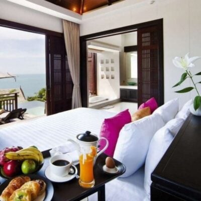 A neatly made bed with pink pillows, a breakfast tray with fruit, coffee, and juice, and an ocean view from a modern, open hotel room with a balcony—creating the ultimate guest room experience.