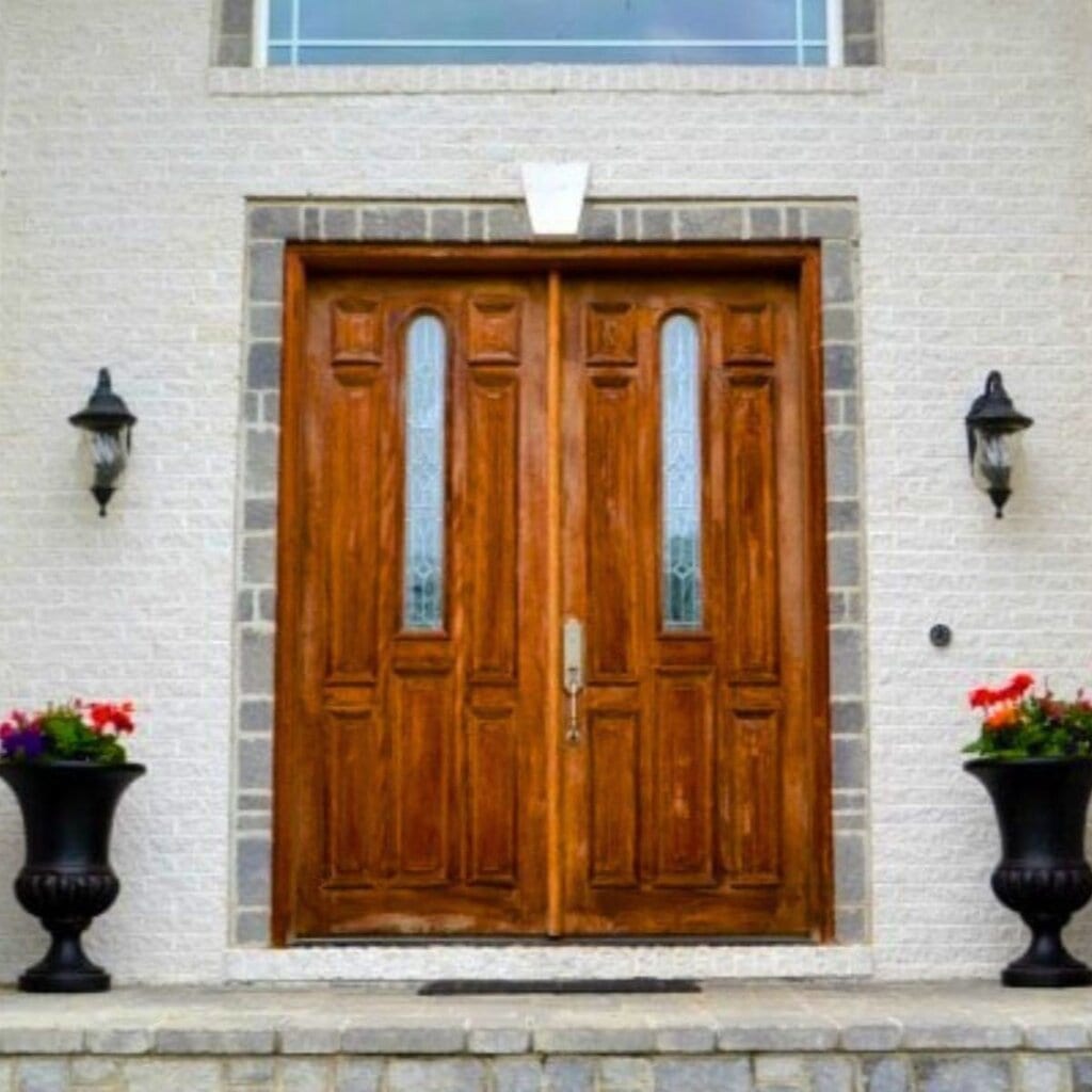 Double wooden front doors with decorative glass panels, flanked by black planters and lantern lights on a white brick exterior—a quick, easy project to enhance your home's curb appeal.