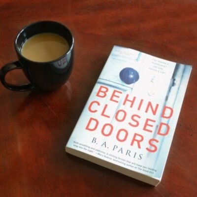 A black mug of coffee sits next to "Behind Closed Doors" by B.A. Paris on a wooden surface, creating a cozy setting for diving into this gripping thriller.