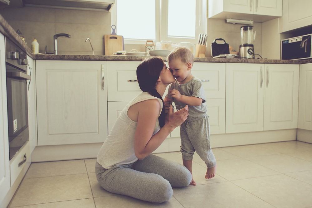 a mom sitting on the kitchen floor giving her toddler son a kiss on his cheek