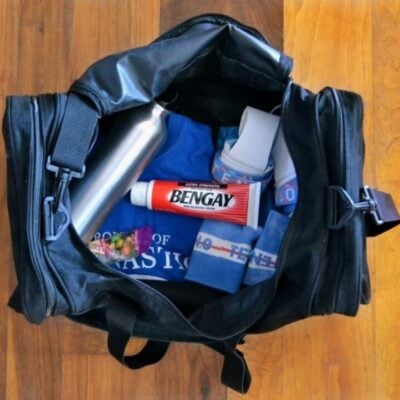 An open black gym bag on a wooden floor contains a water bottle, blue shirt, Bengay for pain relief, athletic tape, resistance band, and colorful pills in a plastic bag—perfect essentials for active teens on the go.