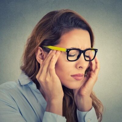 Woman with long hair and glasses holds her temples and closes her eyes, appearing to suffer everyday headaches—she may need to get her eyes checked.