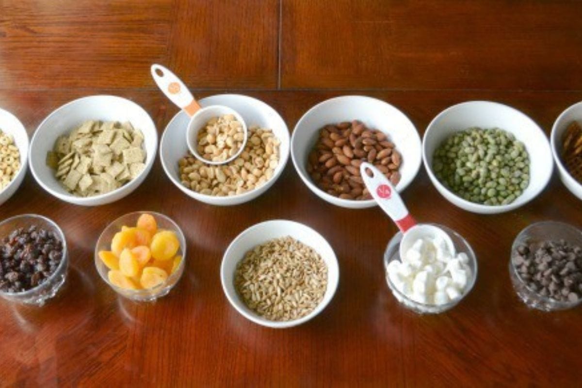 A vibrant trail mix buffet features a variety of nuts, seeds, dried fruits, cereals, and chocolate chips arranged in white bowls and clear cups on a wooden table.