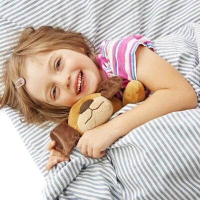 A young child lies on a striped bed, smiling and hugging a stuffed dog toy while partially covered by a striped blanket—ready to start off the morning with energy for back school routines.