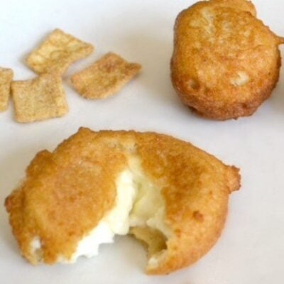 Three square cereal pieces and two Cinnamon Donut Holes with Cream Cheese Filling, one of which is bitten to reveal its creamy white center, are arranged on a plain white surface.
