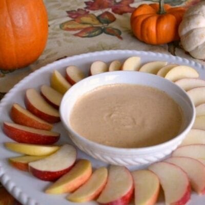 A white plate with sliced apples arranged around a bowl of healthy pumpkin pie dip, set on a table with decorative pumpkins and a fall-themed tablecloth.