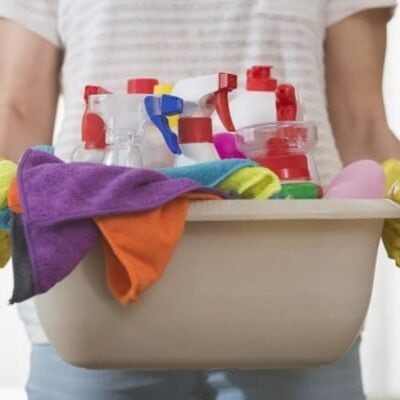 Person holding a beige plastic container filled with cleaning supplies, spray bottles, and colorful cleaning cloths—perfect essentials to keep your home tidy and clean by following a strict, effective declutter routine.
