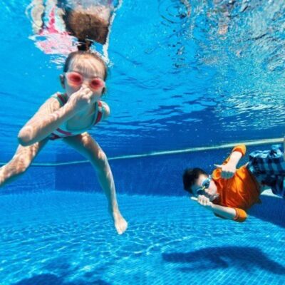 Two children underwater in a pool, both wearing goggles. The girl holds her nose while the boy gives a thumbs up, showing why many wonder—are swimming pools worth it? Blue pool tiles and sunlight shine all around.