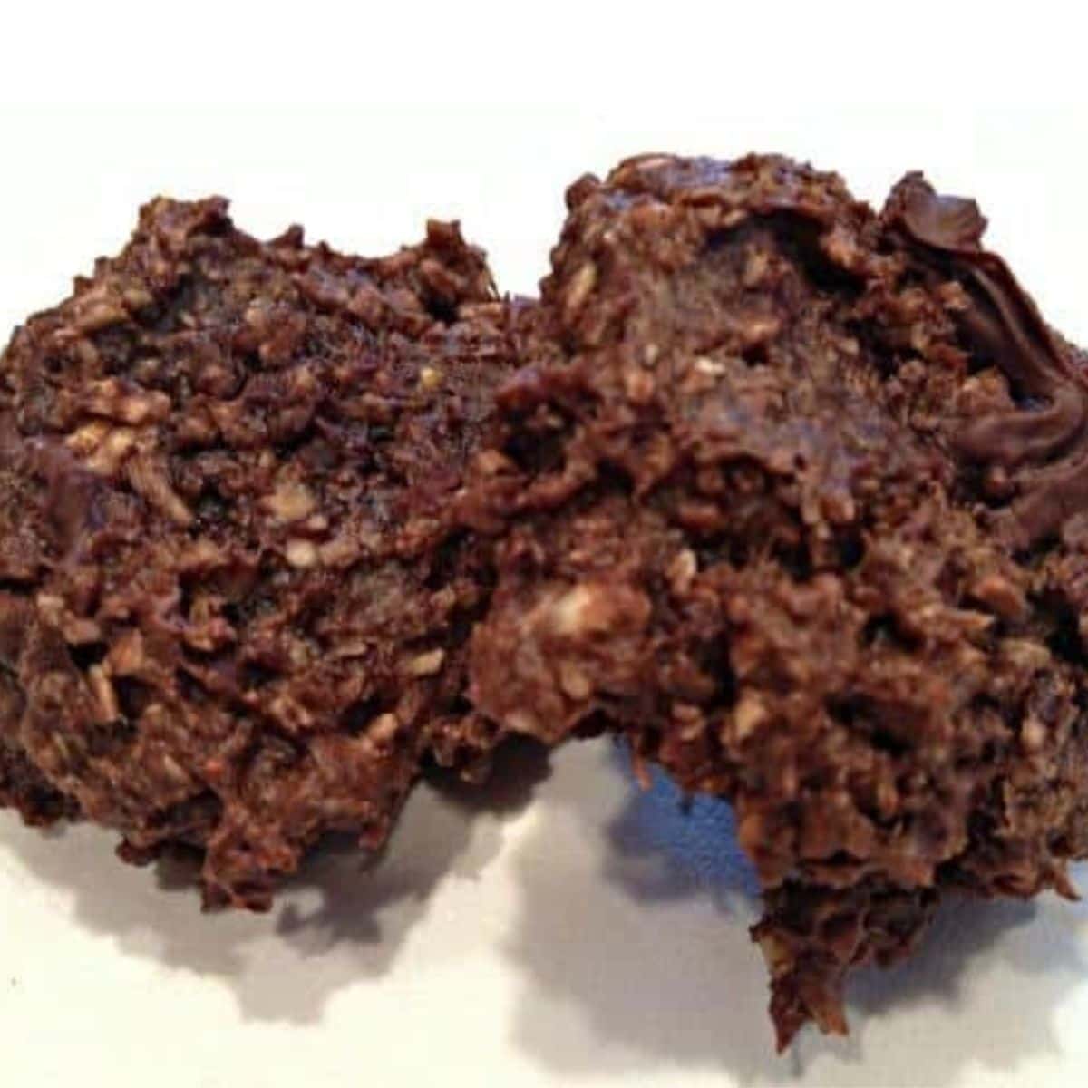 Two no-bake chocolate oatmeal cookies on a white surface, their rough, textured appearance highlighting visible oats and rich chocolate—a delicious example of a simple chocolate protein cookie recipe.