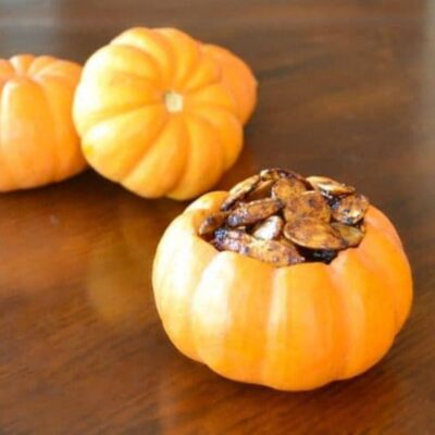 Three small pumpkins rest on a wooden surface, with one in the foreground filled with cinnamon roasted pumpkin seeds.