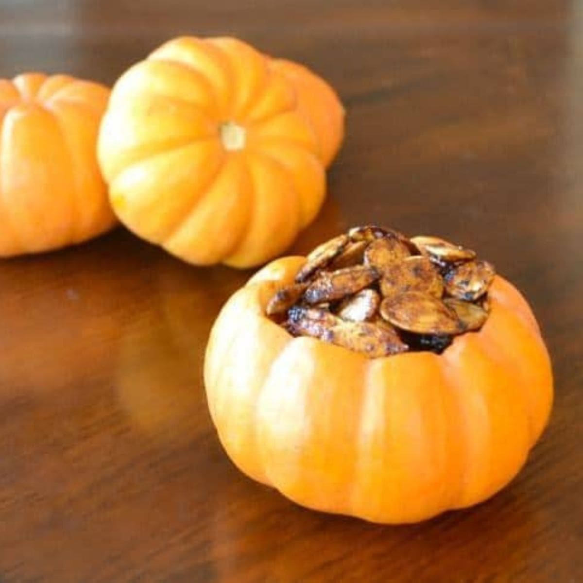 Three small pumpkins rest on a wooden surface, with one in the foreground filled with cinnamon roasted pumpkin seeds.