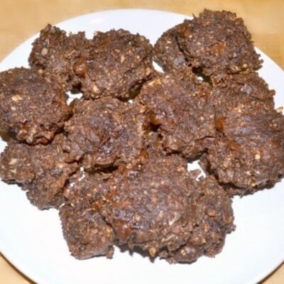 A white plate topped with several dark brown, chunky chocolate protein cookies that appear to contain oats and chocolate.