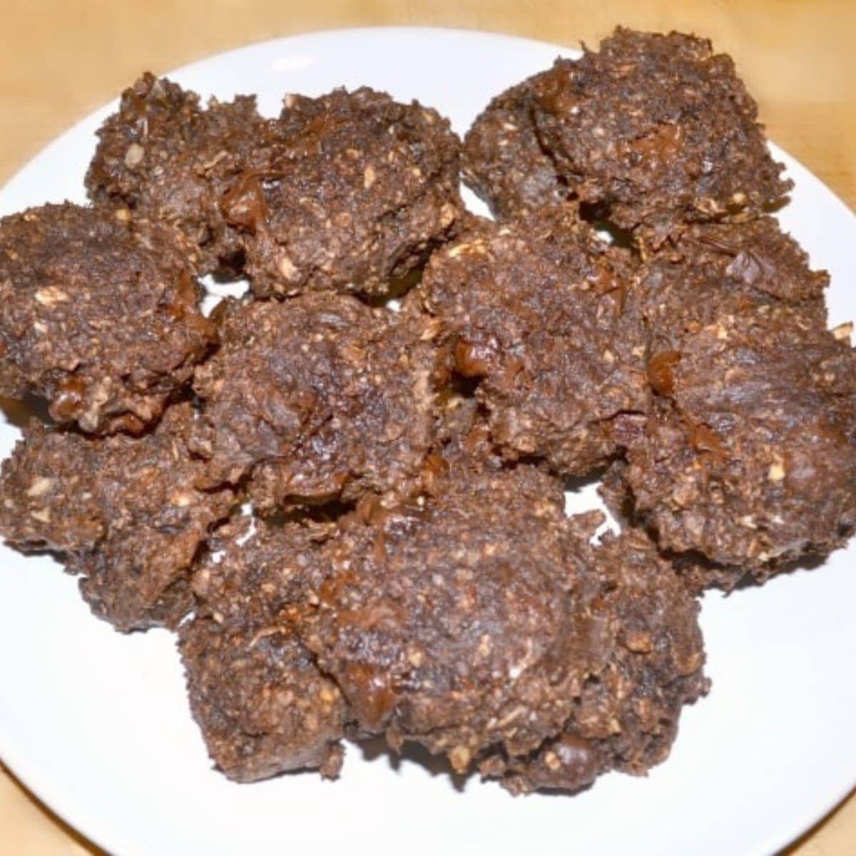 A white plate topped with several dark brown, chunky chocolate protein cookies that appear to contain oats and chocolate.