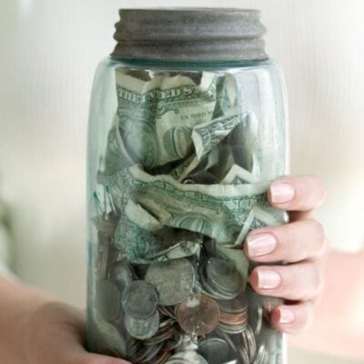 A person holding a clear glass jar filled with assorted U.S. dollar bills and coins, showcasing practical money saving tips in action.