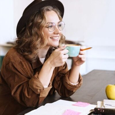 A person wearing glasses and a hat, holding a cup and smiling at a table adorned with notebooks, sticky notes, and an apple—crafting their daily routine to discover new ways to get more done every day.