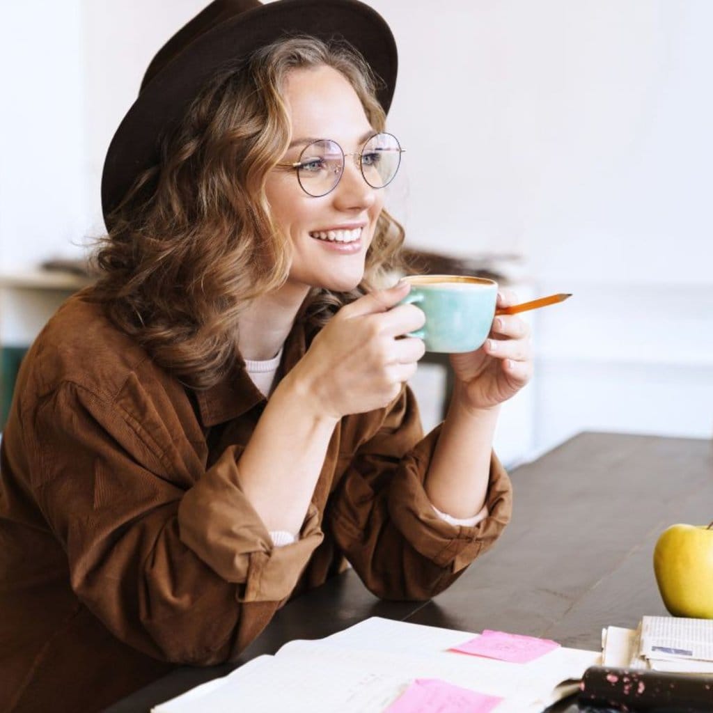 A person wearing glasses and a hat, holding a cup and smiling at a table adorned with notebooks, sticky notes, and an apple—crafting their daily routine to discover new ways to get more done every day.