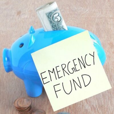 A blue piggy bank with a dollar bill inserted and a sticky note labeled "EMERGENCY FUND," surrounded by scattered coins—a visual reminder of how to create an emergency fund and safeguard your finances.
