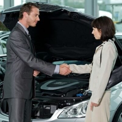 A man and a woman in business attire shake hands in front of a car with its hood open inside a showroom, sealing the best deal on a new car.