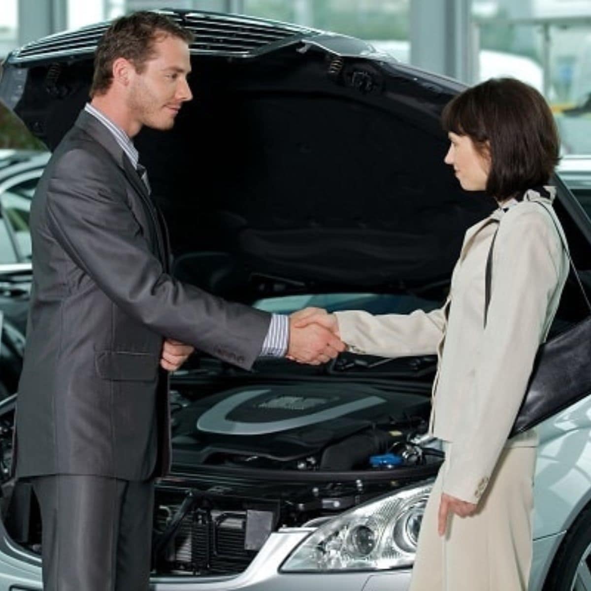 A man and a woman in business attire shake hands in front of a car with its hood open inside a showroom, sealing the best deal on a new car.