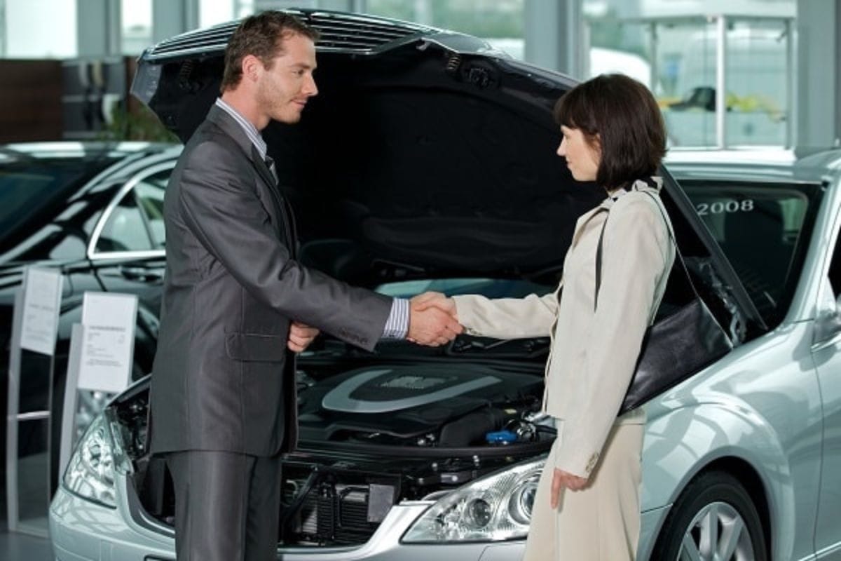 A man in a suit and a woman in a light-colored coat shake hands in front of a silver car with its hood open, sealing the best deal on a new car inside a showroom.