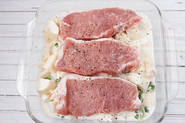 raw pork chops seasoned with salt and pepper on creamy potatoes in a glass baking dish on a white wood table